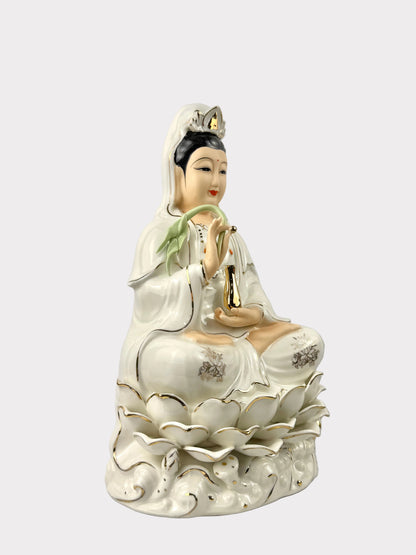 A white porcelain figurine of Guanyin seated on a lotus base, with gold accents and a green wrist scarf, photographed against a plain light gray background.