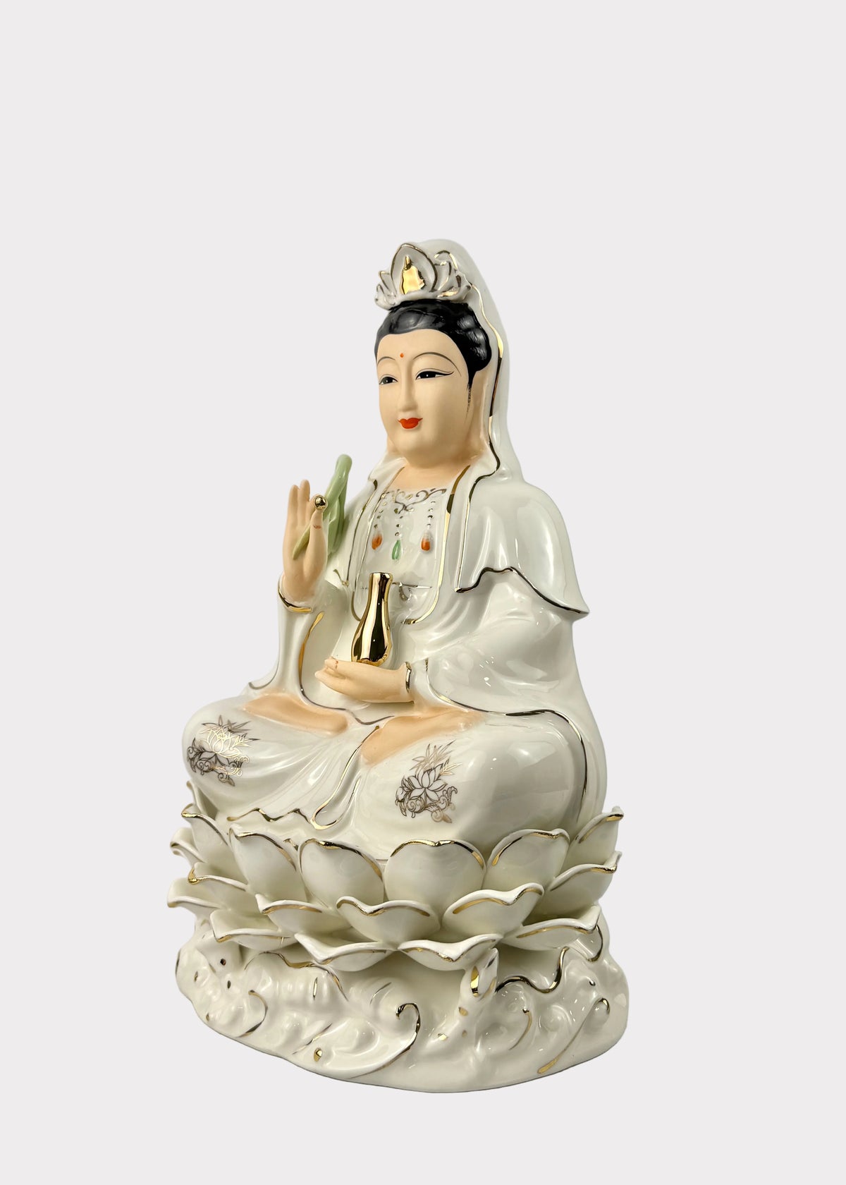 A white porcelain figurine of a seated female deity with black hair, wearing a crown and flowing robes, holding a gold bell, set on a lotus base against a light gray background.