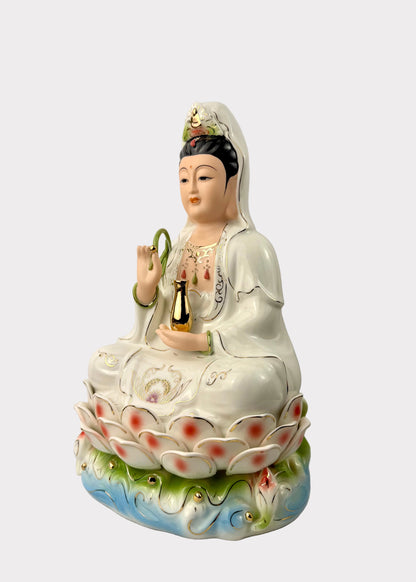 A white porcelain figurine of Guanyin seated on a lotus base, holding a gold bottle and a green ring, with blue and green decorative details on the pedestal, photographed against a plain light gray background.