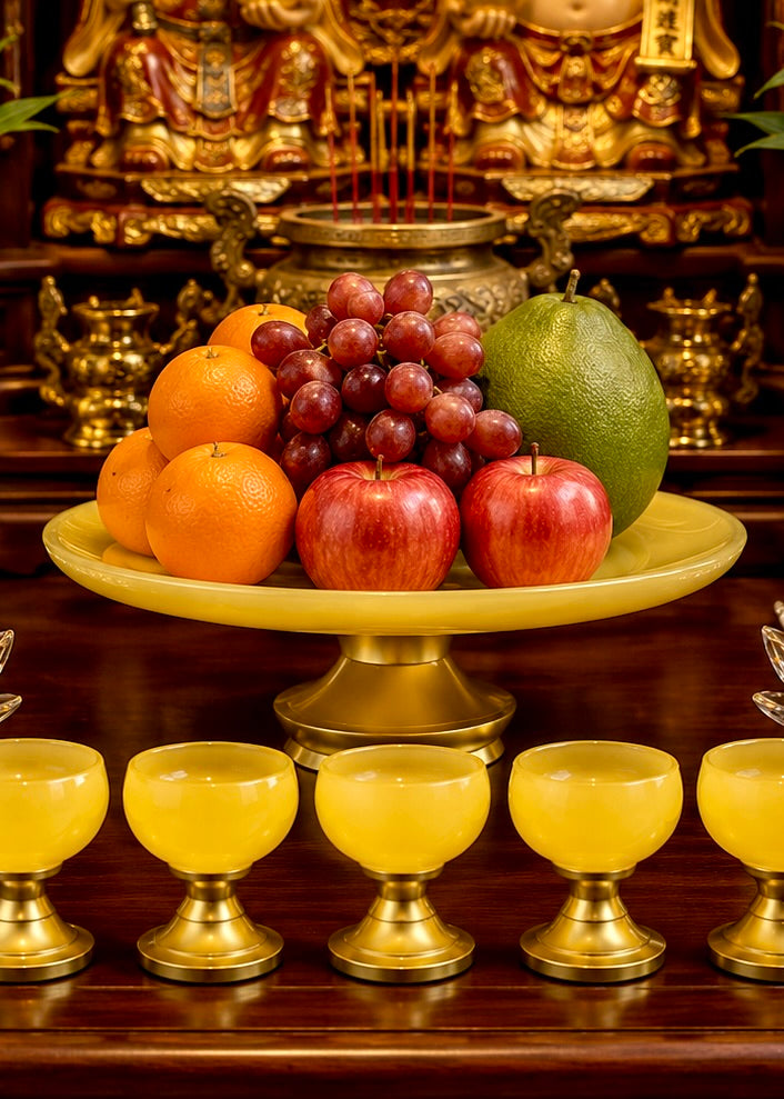 A golden pedestal tray holds a fruit arrangement with oranges, red apples, red grapes, and a green mango, set in front of a decorative gold altar. Five small golden goblets filled with yellow liquid are arranged in a row on a dark wooden surface.