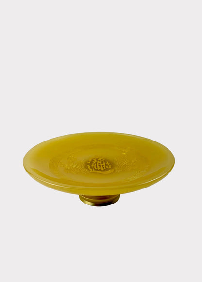 A round, yellow translucent glass pedestal plate with a small gold-colored base, set against a plain light gray background.