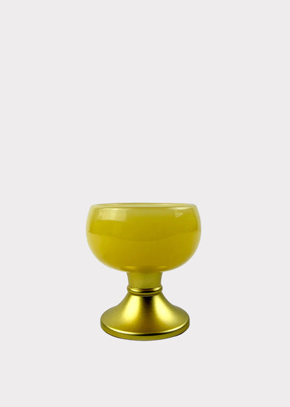 A small yellow glass goblet or candle holder with a metallic gold base, set against a light gray background.