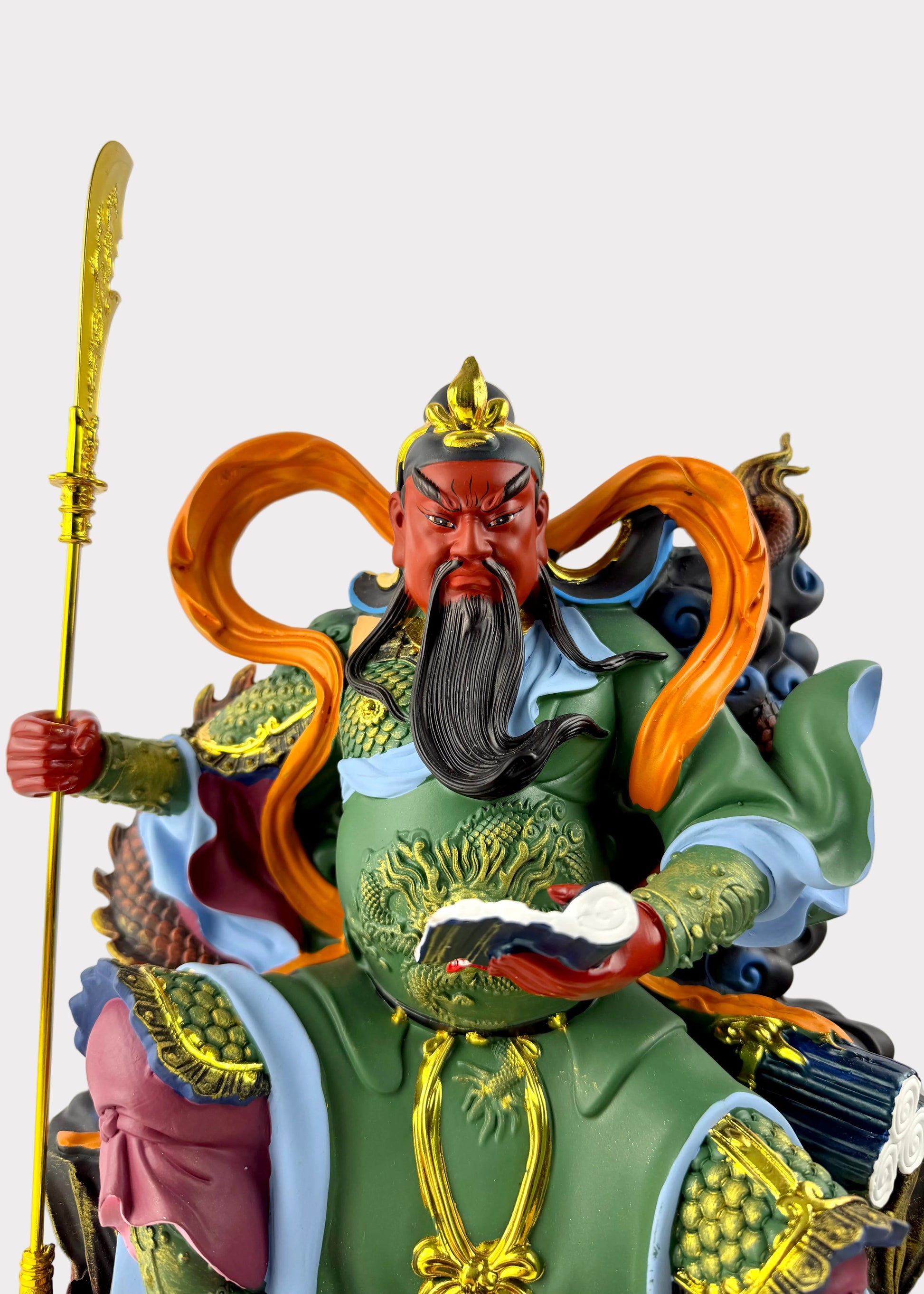 A detailed figurine of a Chinese martial deity, likely Guanyin, depicted with a red face, long black beard, green and gold armor, and flowing orange hair, holding a golden staff against a neutral light gray background.