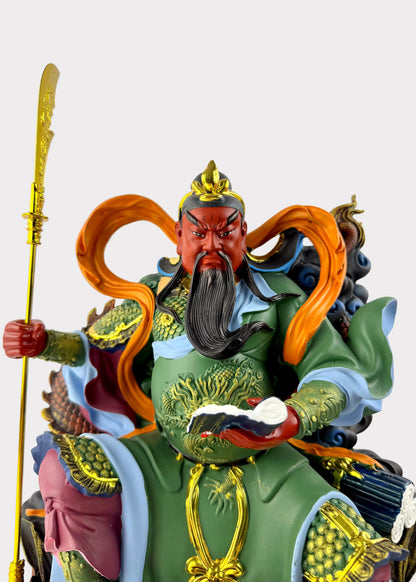 A detailed figurine of a Chinese martial deity, likely Guanyin, depicted with a red face, long black beard, green and gold armor, and flowing orange hair, holding a golden staff against a neutral light gray background.
