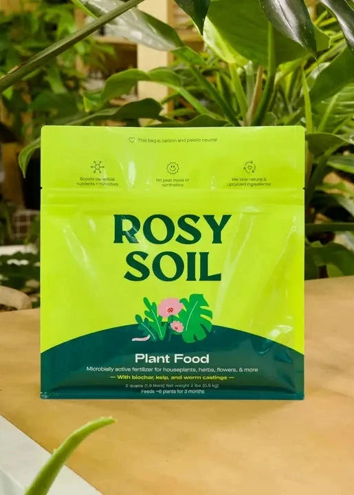 Rosy Soil plant food package on a wooden surface with plants in the background