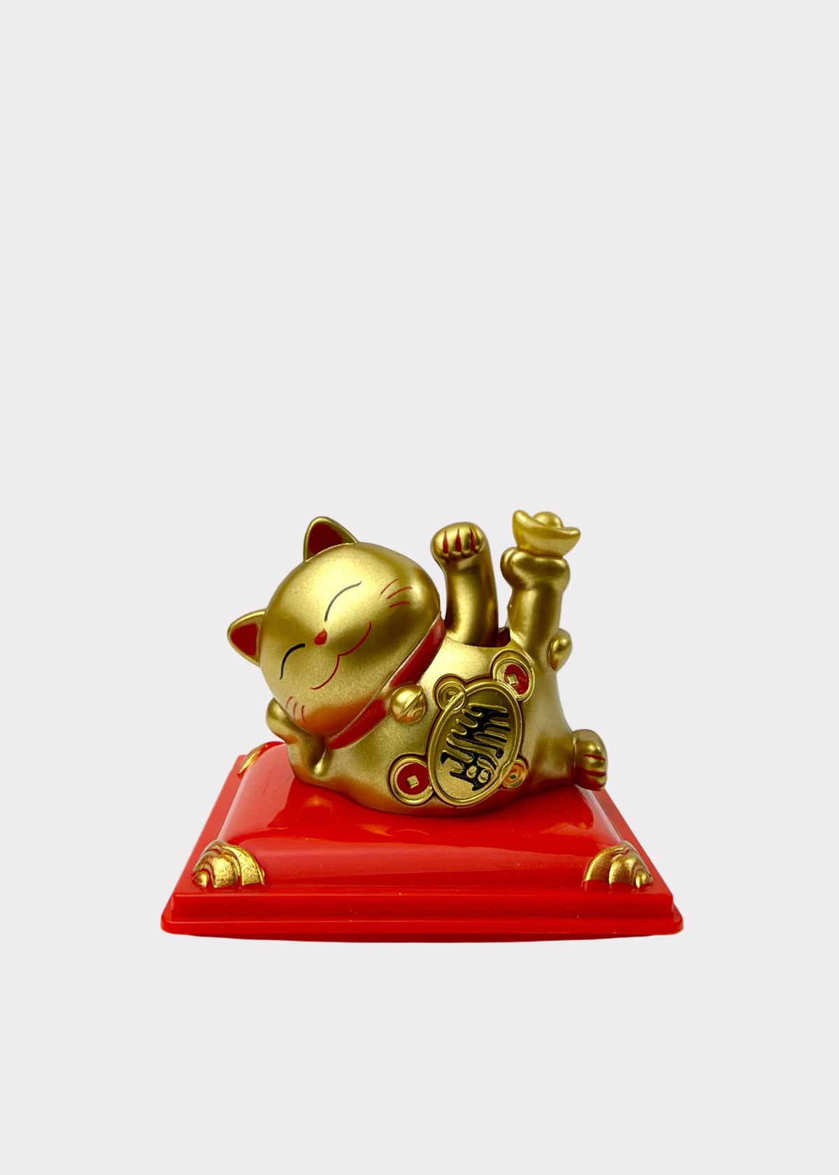 A golden lucky cat figurine lying on its back on a glossy red rectangular base, photographed against a plain light gray background.