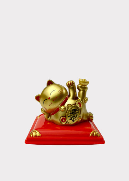 A golden lucky cat figurine lying on its back on a glossy red rectangular base, photographed against a plain light gray background.