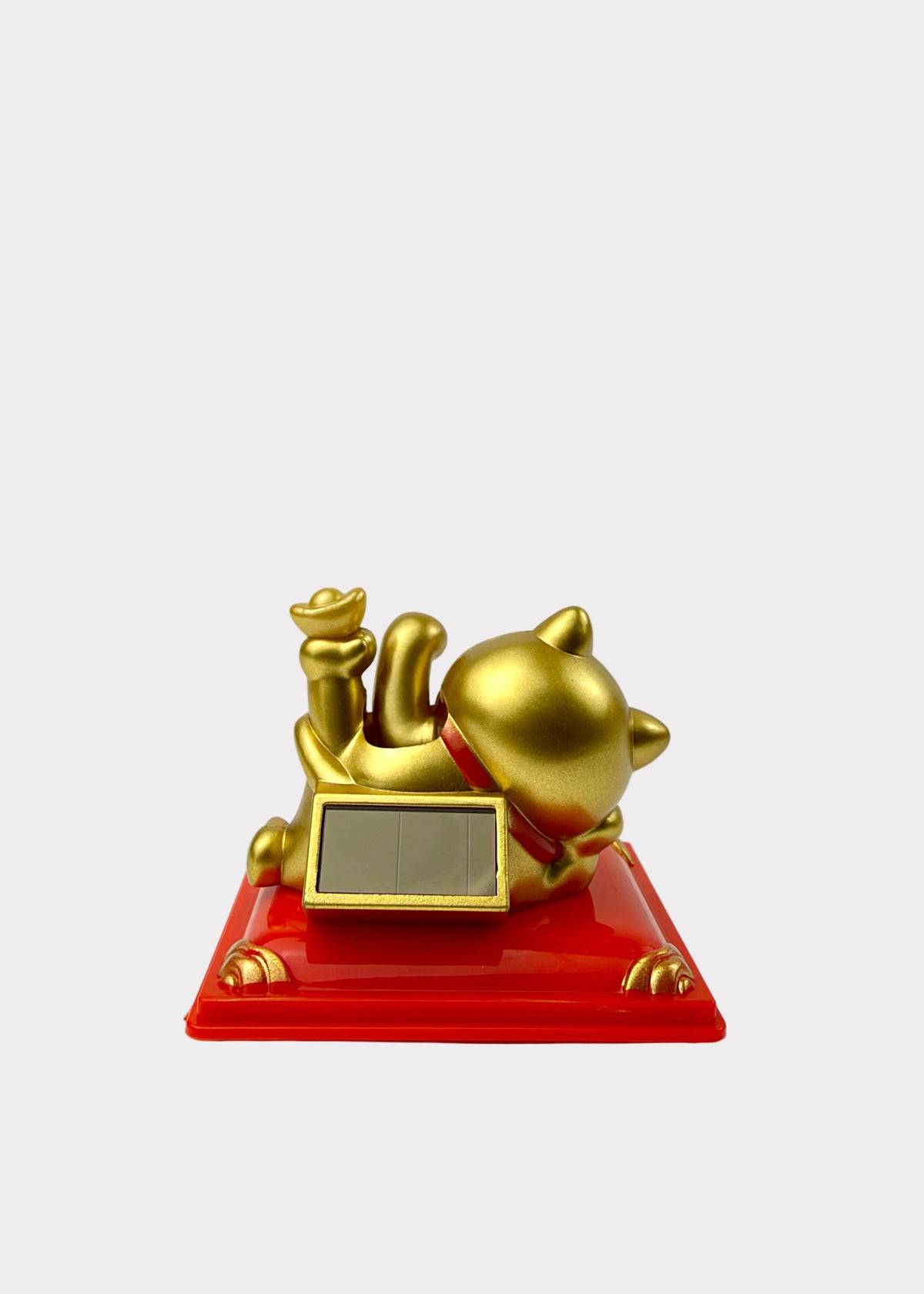 A gold maneki-neko lucky cat figurine lying down on a glossy red rectangular base, with a small blank rectangular plaque on the front and a gold coin held in its front paw, photographed against a plain light gray background.