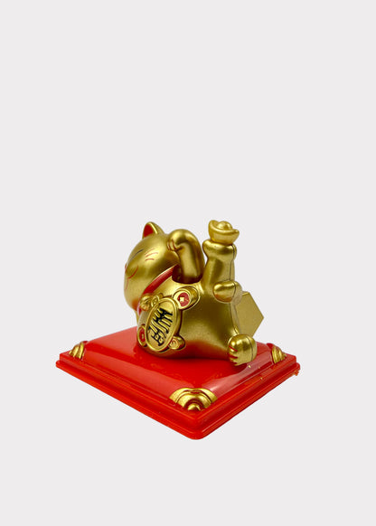 A golden maneki-neko lucky cat figurine holding gold ingots, resting on a glossy red square base with gold corner accents, photographed against a plain white background.