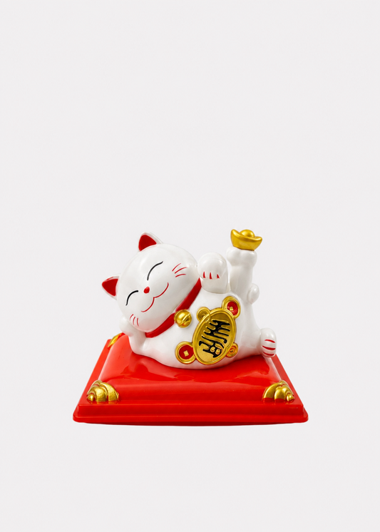 A white ceramic Maneki-neko lucky cat figurine reclining on a red base with gold accents, set against a plain light gray background.