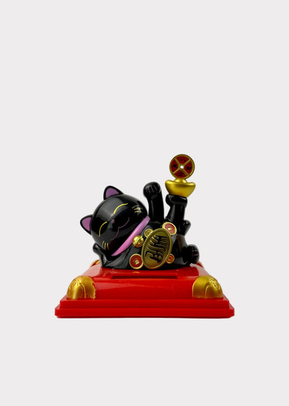 A black lucky cat figurine with gold and pink accents, lying on a red base with gold feet and holding a gold coin and a small gold cup, photographed against a plain light gray background.
