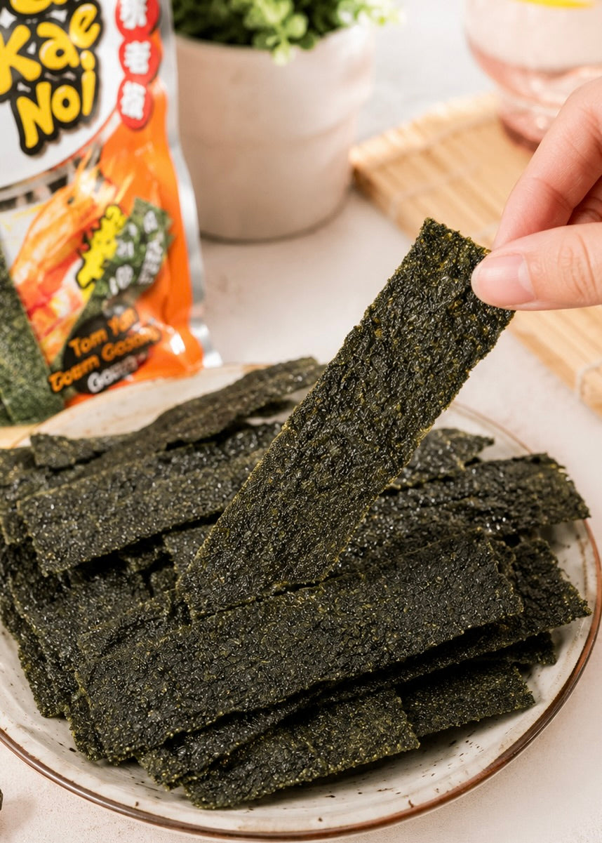 A hand holds a single dark seaweed snack sheet over a plate piled with similar sheets, with a bag of Kake Nori snacks blurred in the background.
