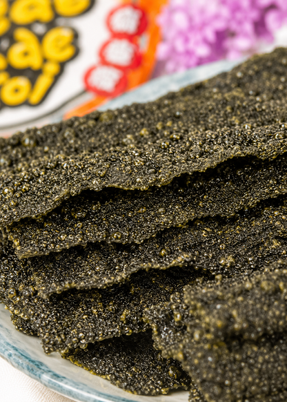 A close-up shot of several dark, crispy seaweed or rice paper snack strips stacked on a plate, with a blurred colorful snack package in the background.